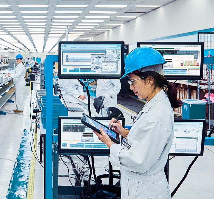 Woman wearing blue hard hat in manufacturing plant holding a tablet.